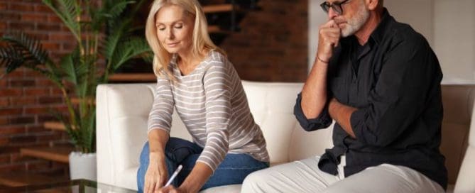 A woman and a man are sitting on a couch in a modern living room. The woman is writing on a piece of paper, while the man watches thoughtfully.