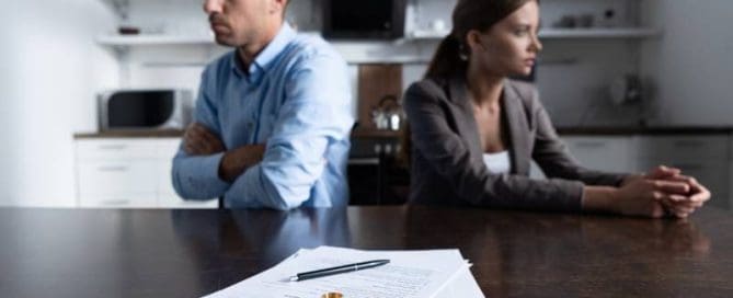 A man and woman sit back to back at a table with divorce papers and wedding rings in the foreground.