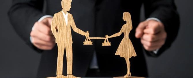 Wooden figures of a man and woman holding scales of justice are positioned in front of a person in a suit.
