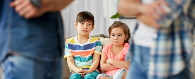 Two children sit on a sofa looking upset, while two adults face each other in the foreground with crossed arms.