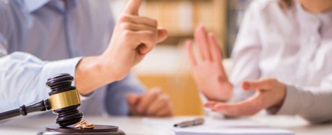 A man and woman sit at a table with papers near a gavel. As the man raises a finger, the woman gestures with open palms, hinting at a discussion or disagreement over custody rights for grandparents.