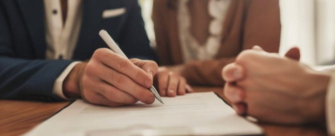 Two people sit at a wooden table as one person signs a document on a clipboard. Their hands are visible, seemingly engaged in a formal agreement or contract review, possibly discussing custody rights for grandparents.