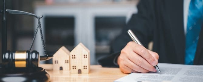 A person in business attire signs a document at a desk with a gavel, scales of justice, and two wooden house models, illustrating what is a prenuptial agreement in California.