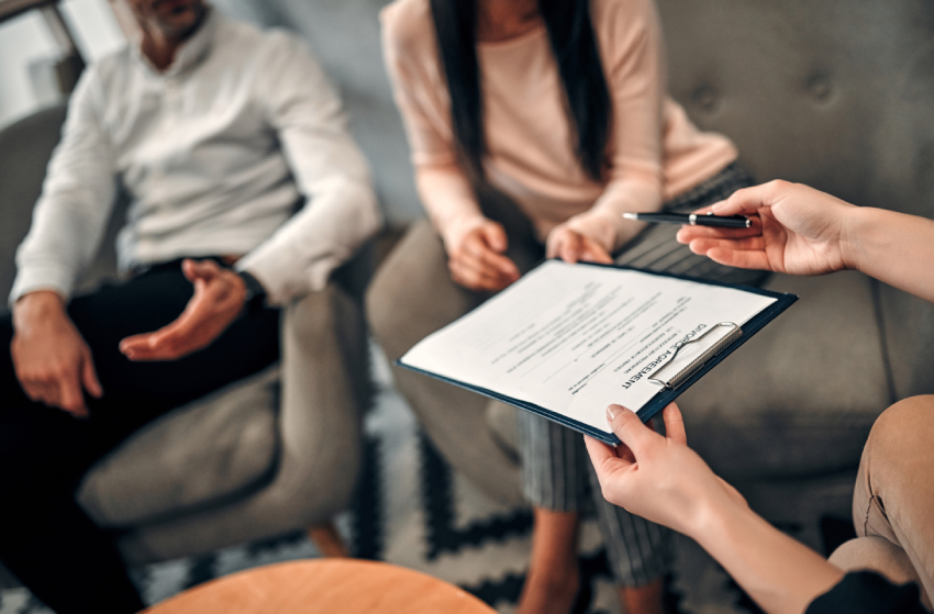 Two people sitting on chairs discuss a military child custody document held on a clipboard by a third person, whose hand gestures with a pen.